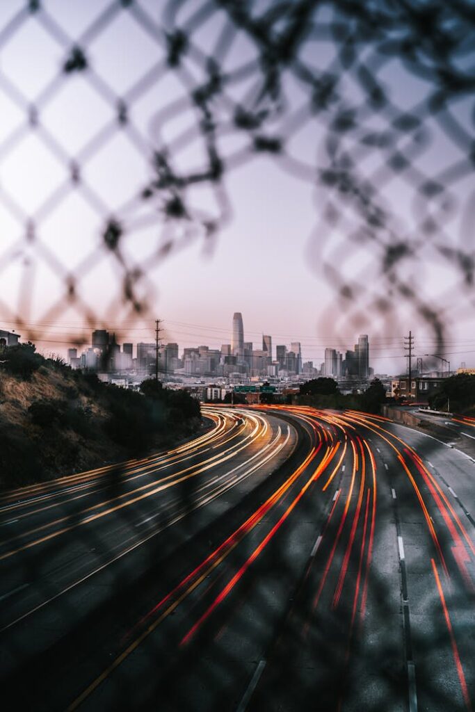 Long exposure of San Francisco highway light trails with city skyline and chain fence foreground.